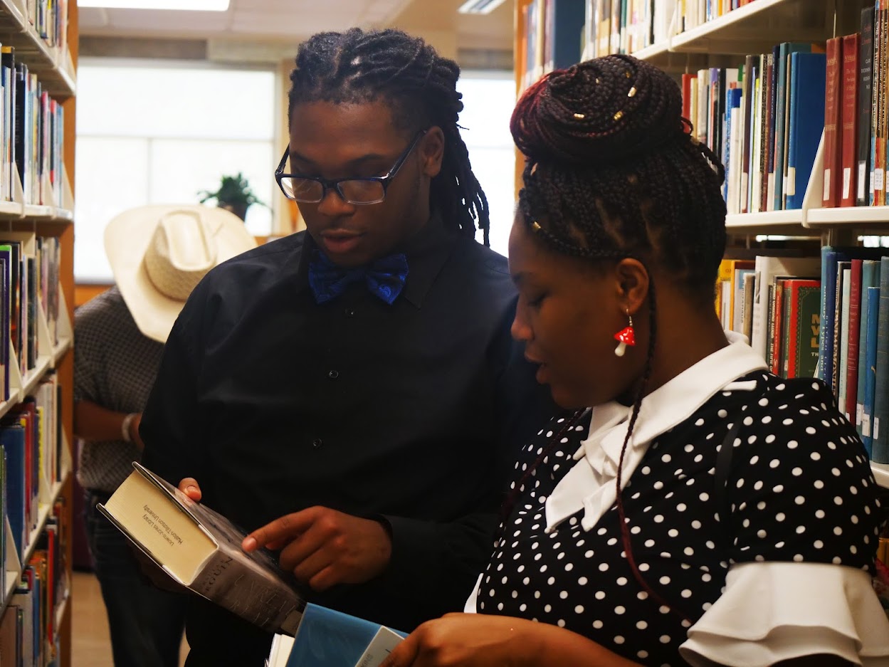 Photo of two HT students discussing books in the library shelves.