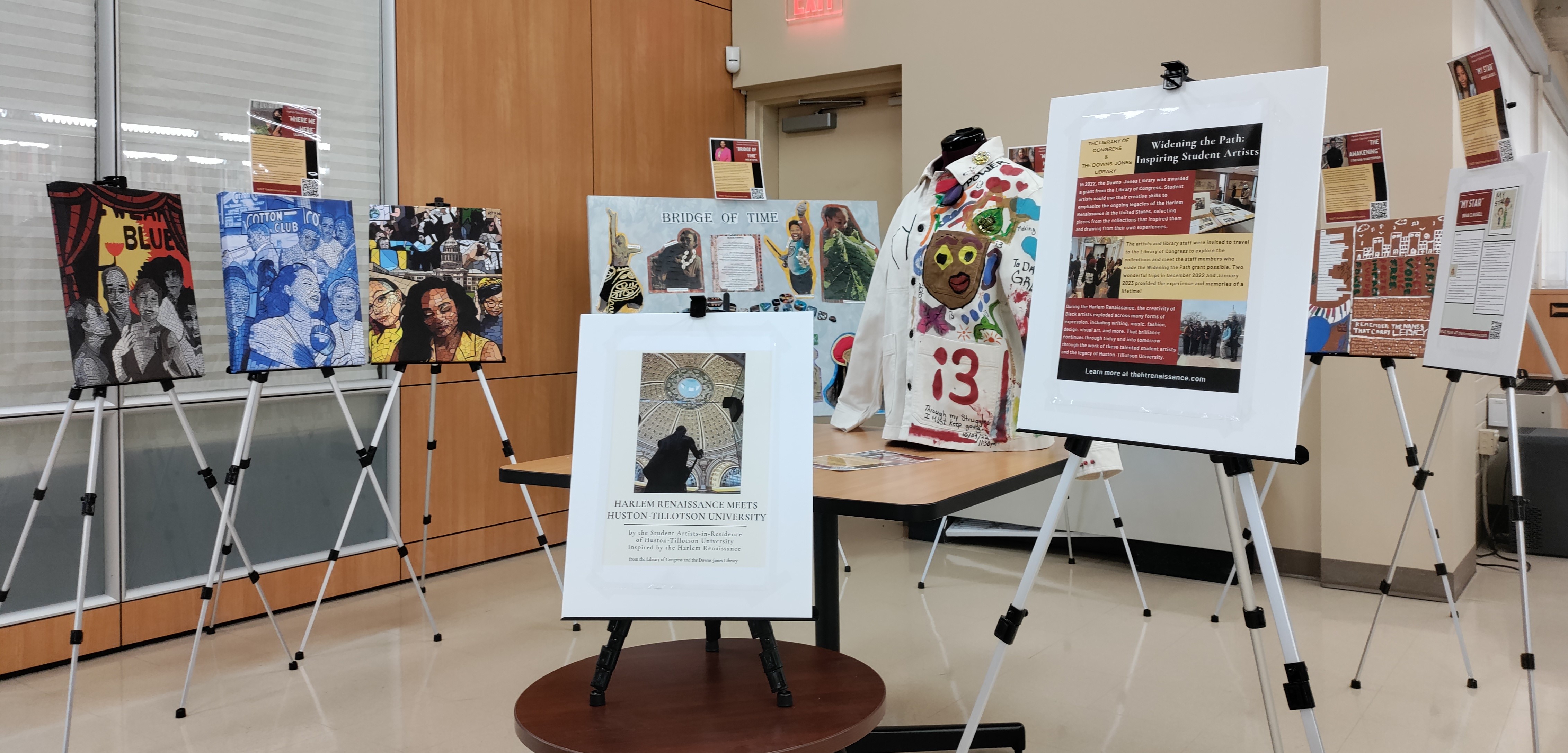 Photograph of an art exhibit in the library. Multimedia artworks are displayed on easels surrounding a central table.